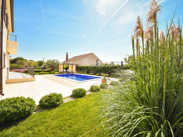 A serene outdoor area features a well-maintained lawn bordered by ornamental grasses and shrubs. A clear blue swimming pool reflects sunlight, accompanied by comfortable lounge seating. A shaded area near the pool invites relaxation, while a light-colored building completes the peaceful landscape.