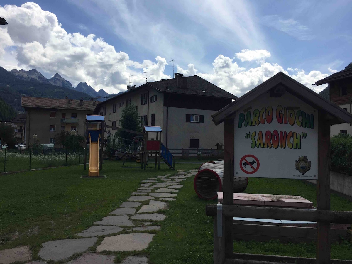 A children's playground is visible, featuring wooden play structures, including slides and climbing areas. A stone pathway leads through the grassy space, with mountains in the background under a partly cloudy sky. A sign indicates the playground's name, 'Parco Giochi Saronch'.