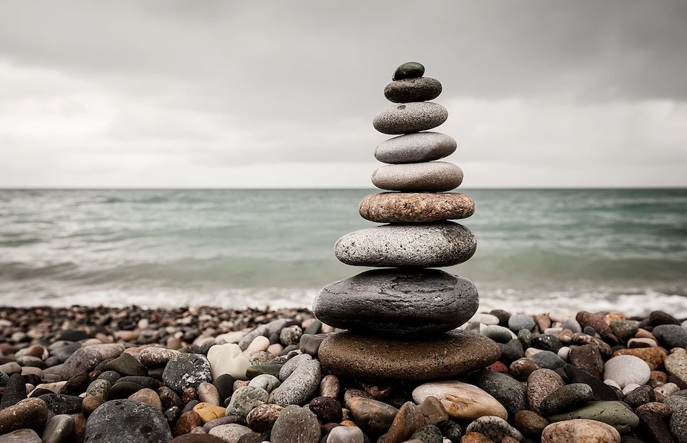 A stack of stones is balanced atop one another on a pebble-strewn beach, with gentle waves lapping at the shore in the background. The scene is set under a cloudy sky, adding a serene quality to the coastal landscape.