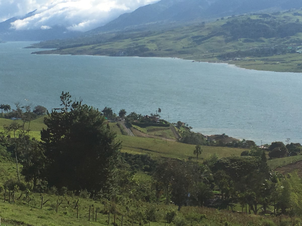 A serene view of Lake Calima is displayed, surrounded by rolling green hills and tropical forests. The landscape is complemented by gentle waves on the water's surface and a distant shoreline dotted with trees and vegetation.