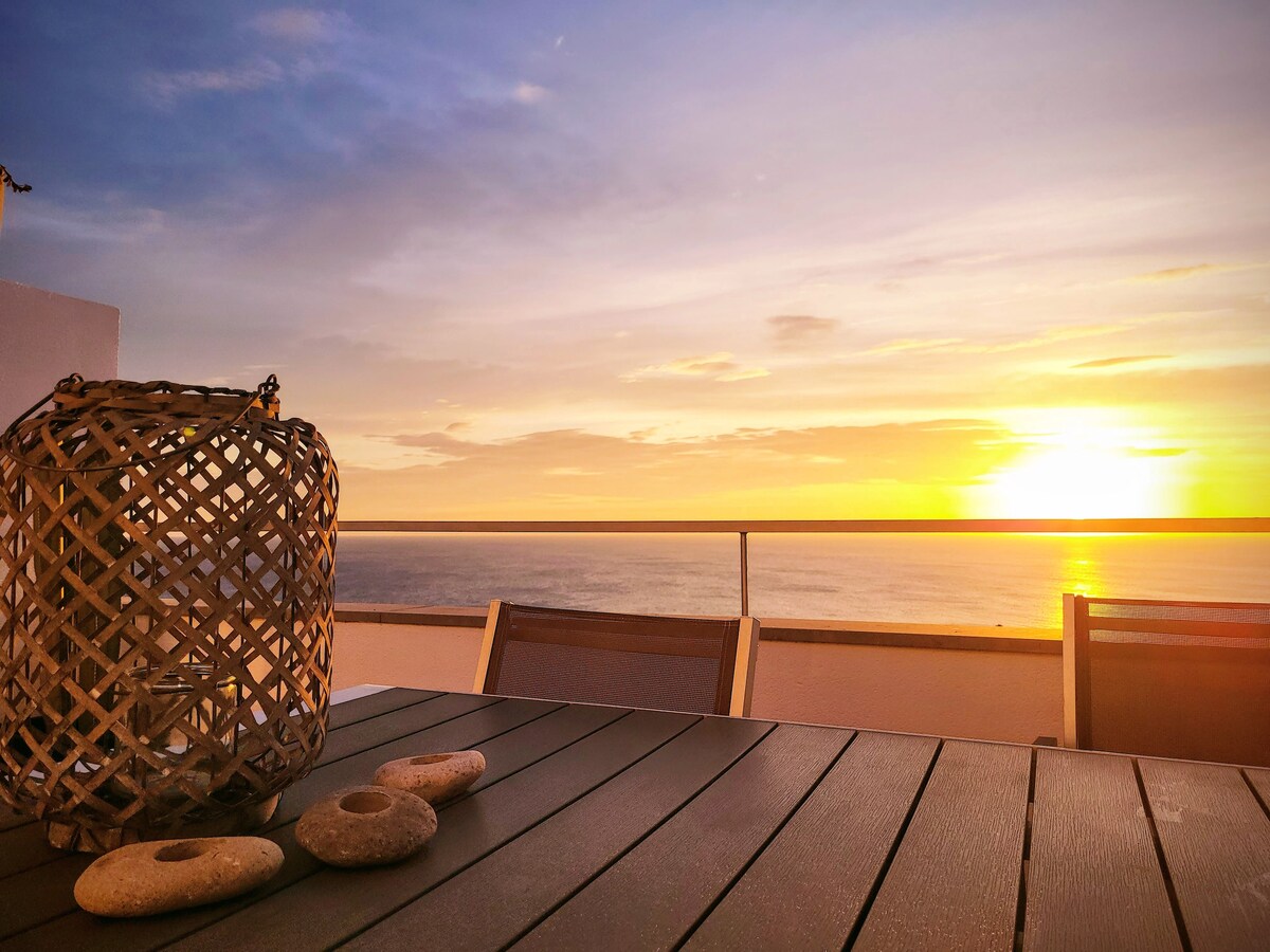 A terrace table is set with a woven lantern and several smooth stones, facing a stunning view of the sunset over the ocean. Soft orange and yellow hues fill the sky, creating a serene backdrop for outdoor gatherings.