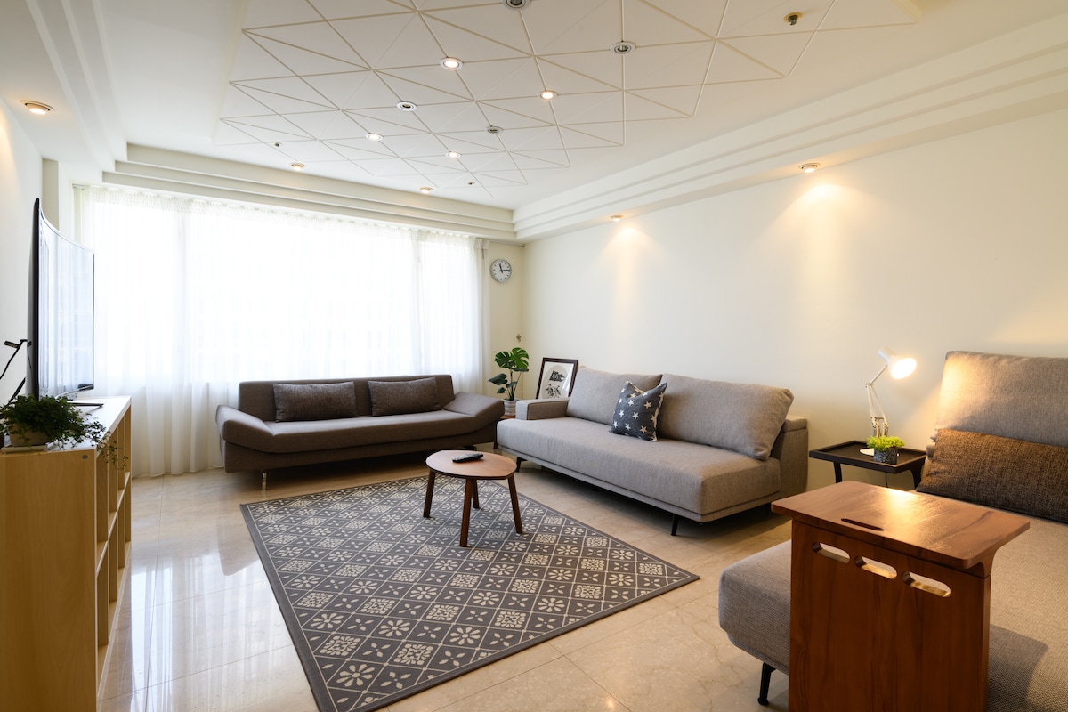 A spacious living area features a large, light-colored sectional sofa and a smaller, sleek armchair. A circular wooden table sits on a patterned rug, with soft natural light filtering through the large window dressed in sheer curtains.