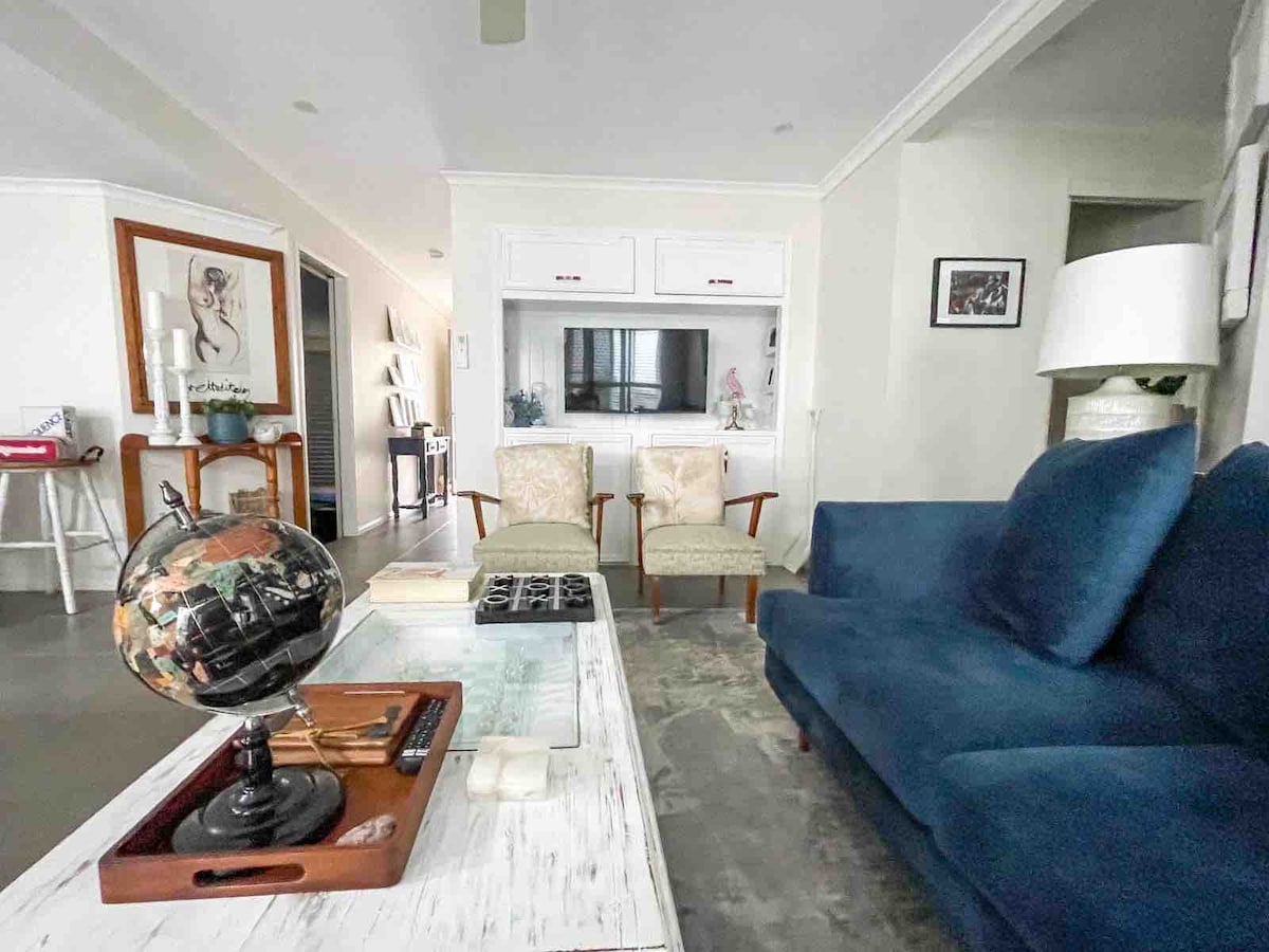 A living area features a blue sofa paired with two armchairs. A coffee table holds a decorative globe and a tray. A television is mounted on the wall within a white cabinetry unit. Light illuminates the space softly, enhancing the neutral tones throughout.
