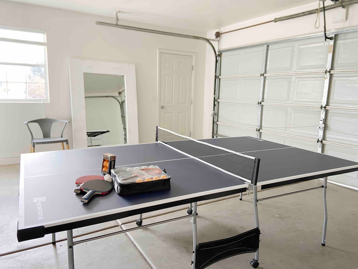 A recreational space is presented with two ping pong tables set up, complete with paddles and a box of balls. A mirror leans against the wall, and a single chair sits nearby. Natural light fills the area through a large window.