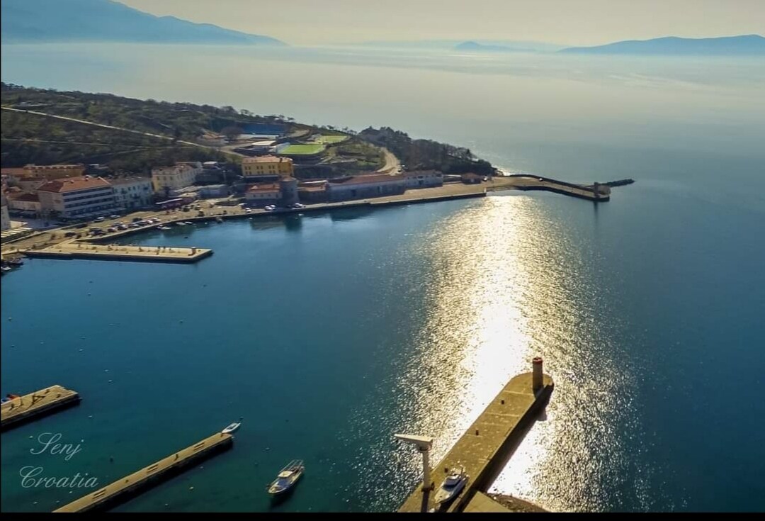 The image captures a serene coastal view of a harbor in Senj, Croatia. Sunlight reflects off the calm blue waters, creating a shimmering pathway. Distant hills and buildings line the shoreline, while boats are docked at the piers, contributing to the tranquil maritime atmosphere.
