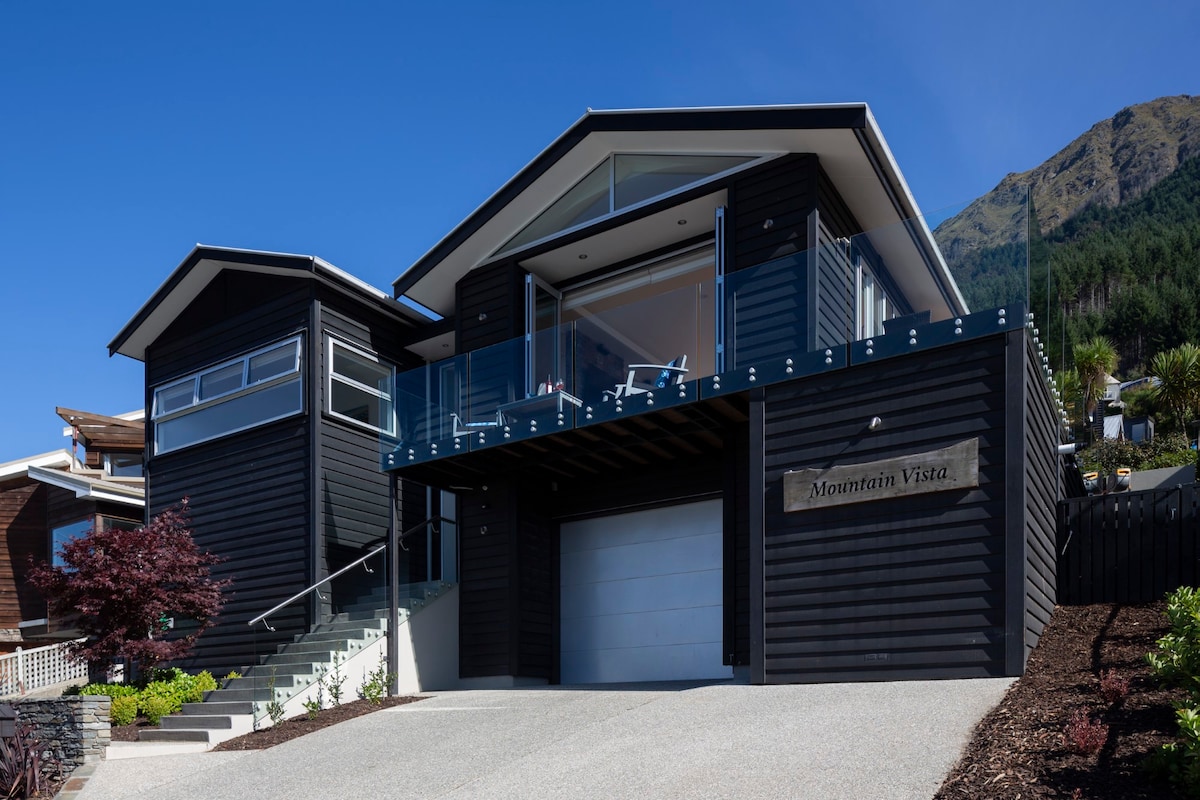 The modern exterior of Mountain Vista is presented, showcasing a sleek black wooden façade and large glass elements. A spacious entryway leads to the home, framed by well-maintained landscaping and vibrant plants. The elevated position allows for expansive views of the surrounding mountains.