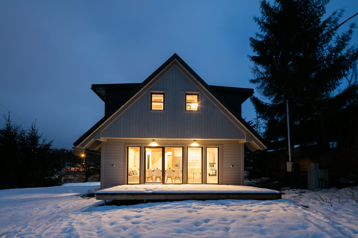 A modern wooden cabin is illuminated at dusk, showcasing large windows that allow a warm glow to emanate from the interior. Fresh snow blankets the surrounding landscape, with tall trees providing a serene backdrop.