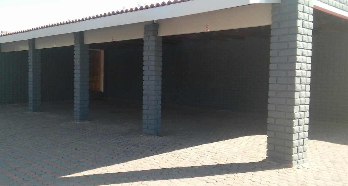 A covered parking area is shown with several pillars supporting the roof. The paved floor exhibits a patterned design, while the walls are painted in a solid dark hue. The entrance features a wooden door, providing access to the adjacent space.