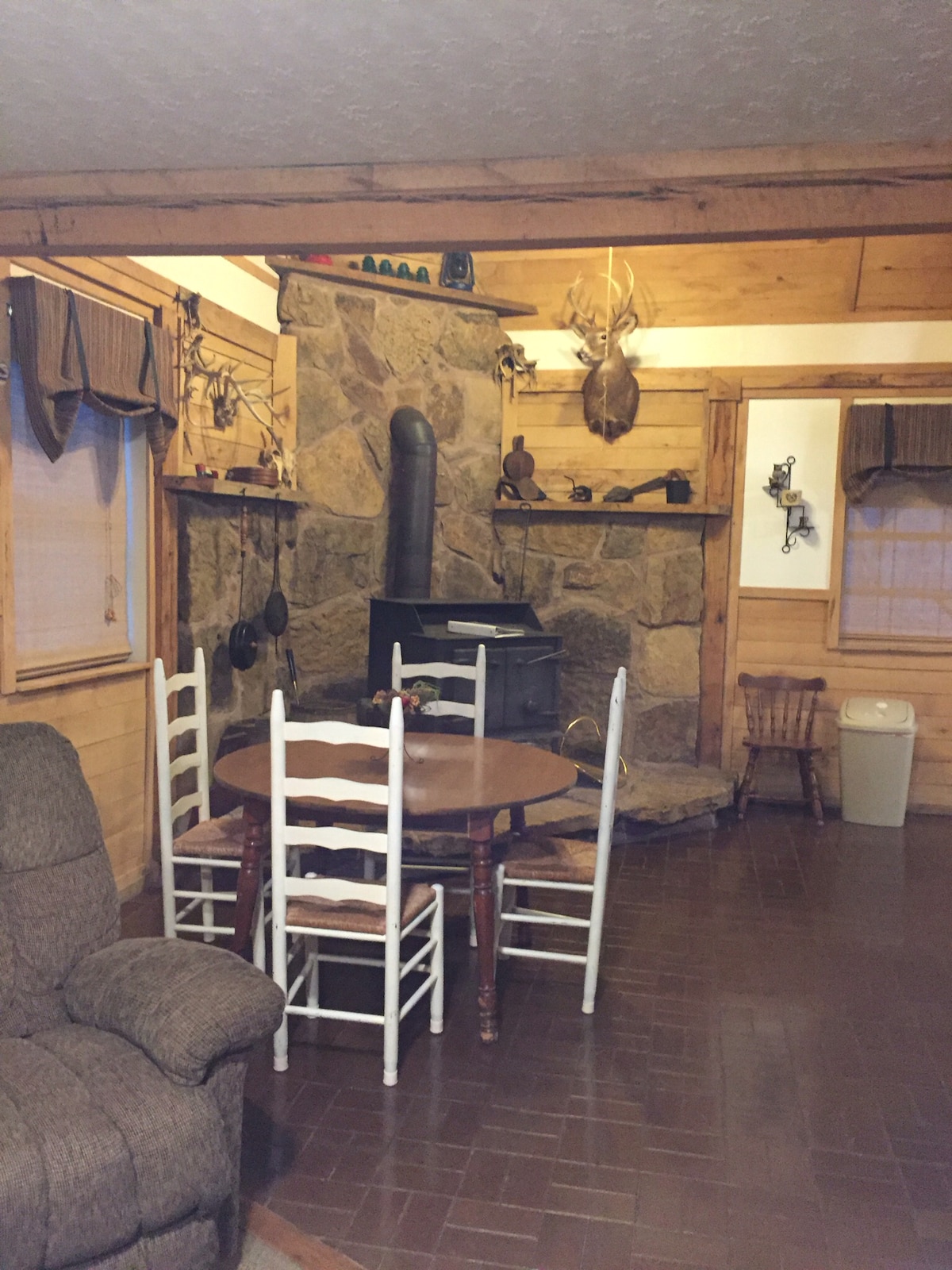 A rustic living space features a stone fireplace as its focal point, with wooden walls and adorned with nature-inspired decor. A round table surrounded by white chairs offers seating for meals. Nearby, a cozy armchair complements the warm ambiance of the cabin.