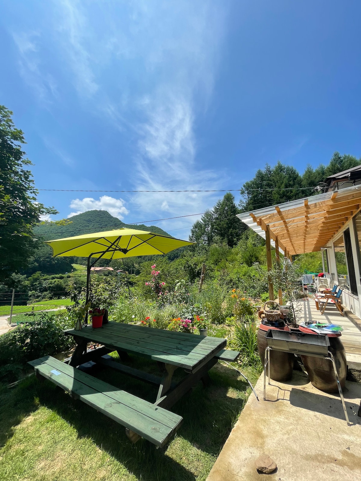 A sunlit outdoor area features a picnic table shaded by a vibrant yellow umbrella. Surrounding flowers and greenery create a natural setting, with a mountain view visible in the background. A small deck with seating is seen to the right, enhancing the inviting atmosphere.
