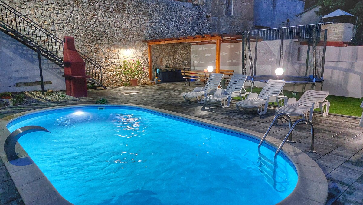 A private outdoor area features a sparkling blue pool surrounded by several sun loungers and a shaded seating area. Soft lighting enhances the ambiance, while a stone wall and greenery provide a natural backdrop, offering a sense of seclusion and relaxation.