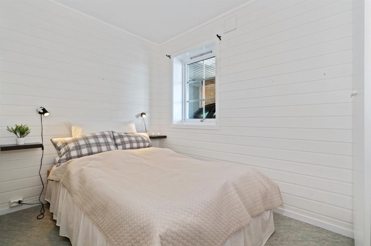 A cozy bedroom features a spacious bed dressed in neutral linens, with decorative pillows. Two wall-mounted lamps provide task lighting beside the bed. A small plant sits on a shelf, and a window offers natural light, enhancing the room's airy feel.