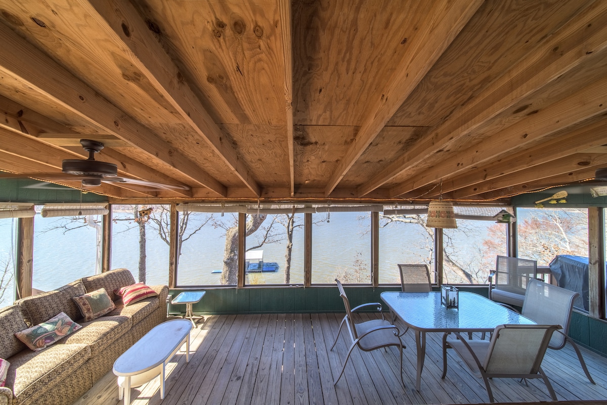 A spacious screened-in porch features wooden beams overhead and various seating options, including a patterned sofa and a dining table. Large windows provide views of the lake and surrounding trees, allowing natural light to flood the space.