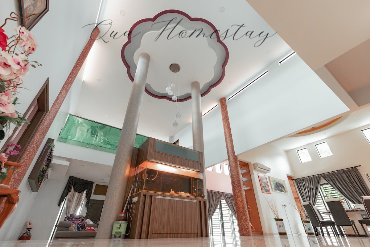 The spacious foyer of the homestay features high ceilings and tall columns, creating an open and airy ambiance. Natural light floods the area through large windows, while a fish tank adds a touch of tranquility. Decorative elements enhance the elegant design.
