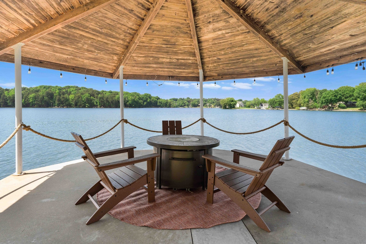 A covered gazebo with a wooden ceiling offers a central firepit surrounded by four wooden chairs. The serene view of Lake Norman stretches out in the background, framed by lush greenery along the shoreline.