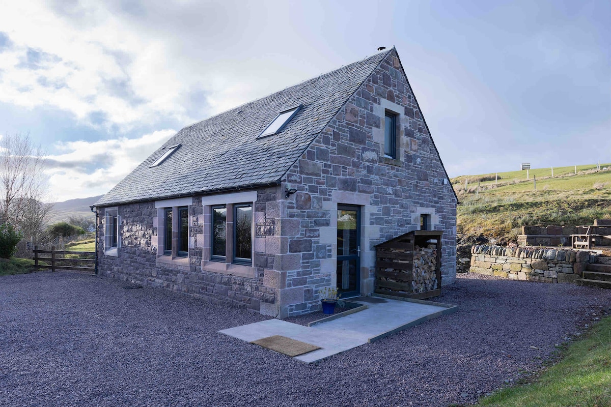 The stone cottage features a sloped roof and large windows that allow natural light to illuminate the interior. A gravel driveway leads to the entrance, with a welcoming entryway visible. The surrounding landscape includes grassy hills, enhancing the cottage's serene setting.