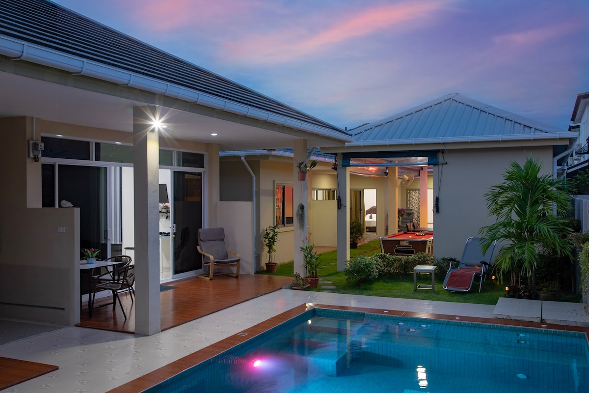 The outdoor area features a well-maintained swimming pool reflecting hues of the sunset, surrounded by a grassy landscape. A pathway leads to a cozy seating area with a table and chairs, while two residential structures are visible in the background with ample windows, creating an inviting ambiance.