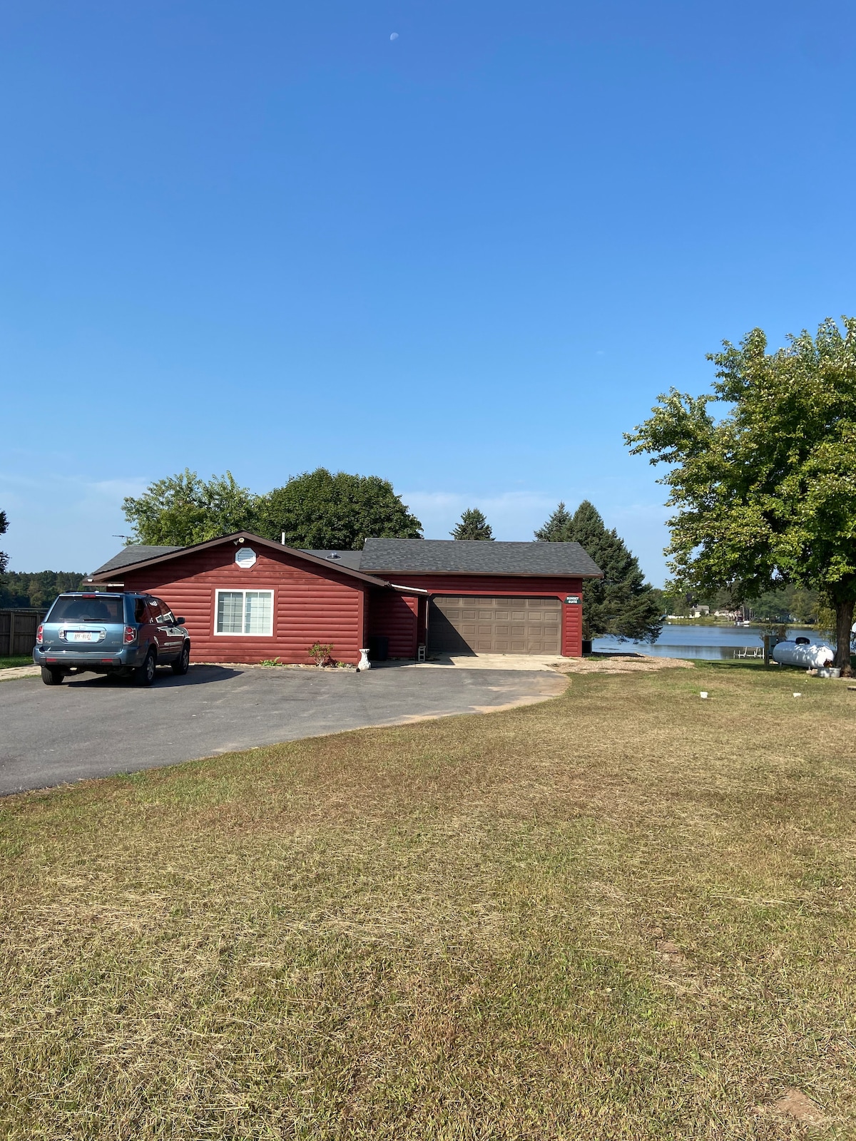 A red duplex is shown with a garage and a paved driveway, situated on a grassy area. Trees are visible nearby, and the water can be seen in the background under a clear blue sky.