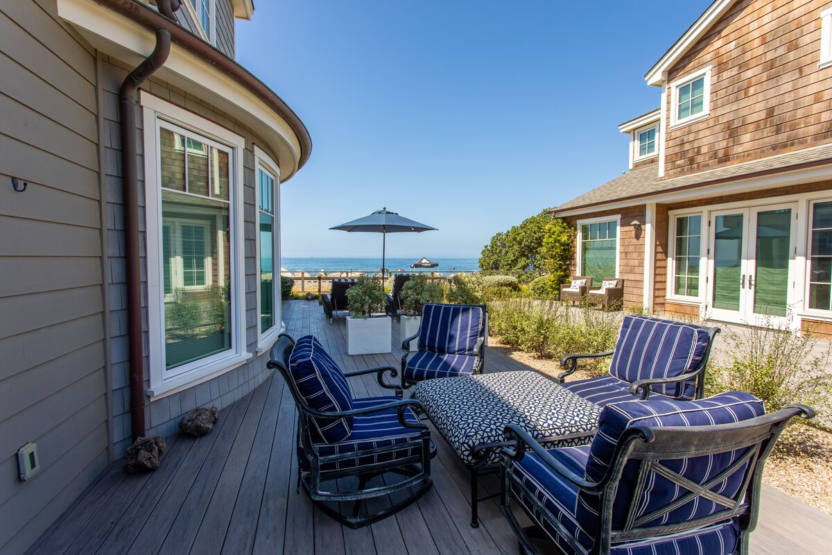 An outdoor seating area features four chairs and a table, all designed with navy and white striped cushions. An umbrella provides shade while a view of the sea is visible in the background, framed by lush greenery and nearby buildings.