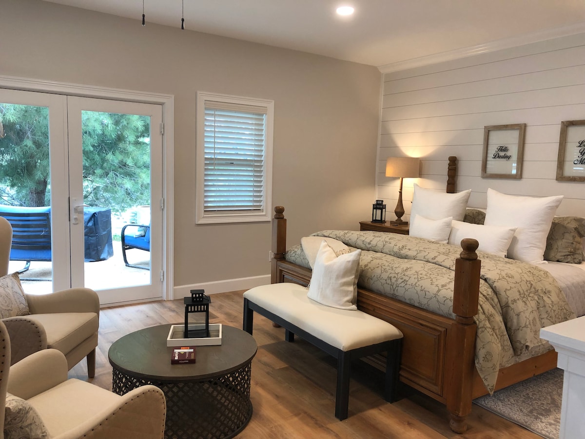 The guest suite features a spacious bed adorned with decorative pillows and a patterned coverlet. A cozy seating area includes two armchairs and a circular coffee table. French doors provide access to a private outdoor space, allowing natural light to enhance the inviting environment.