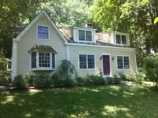 A charming yellow Cape Cod-style house is set amidst greenery, featuring a distinctive front door in deep purple. Multiple windows allow for ample natural light, while lush landscaping provides a serene, inviting environment.