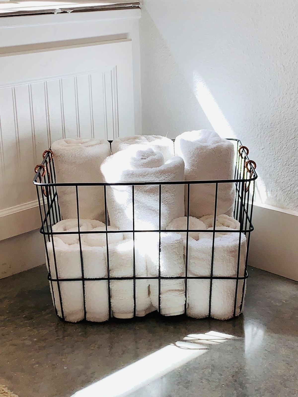 A wire basket contains neatly rolled white towels, arranged upright and evenly stacked. The light from a nearby window casts a gentle shadow on the clean surface below, enhancing the minimalist aesthetic of the space.