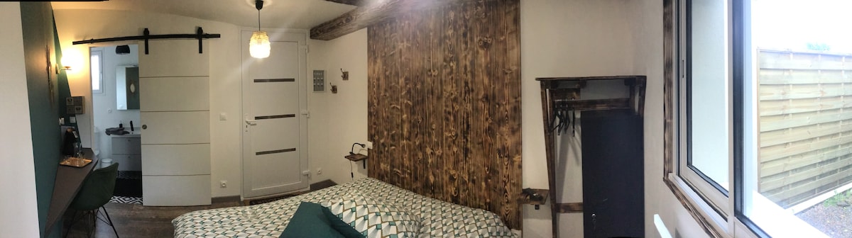 A serene bedroom features wooden wall accents and a spacious bed adorned with patterned bedding. A desk with a chair is positioned near the window, offering natural light. Access to a bathroom is visible through a doorway, enhancing the room's functional layout.