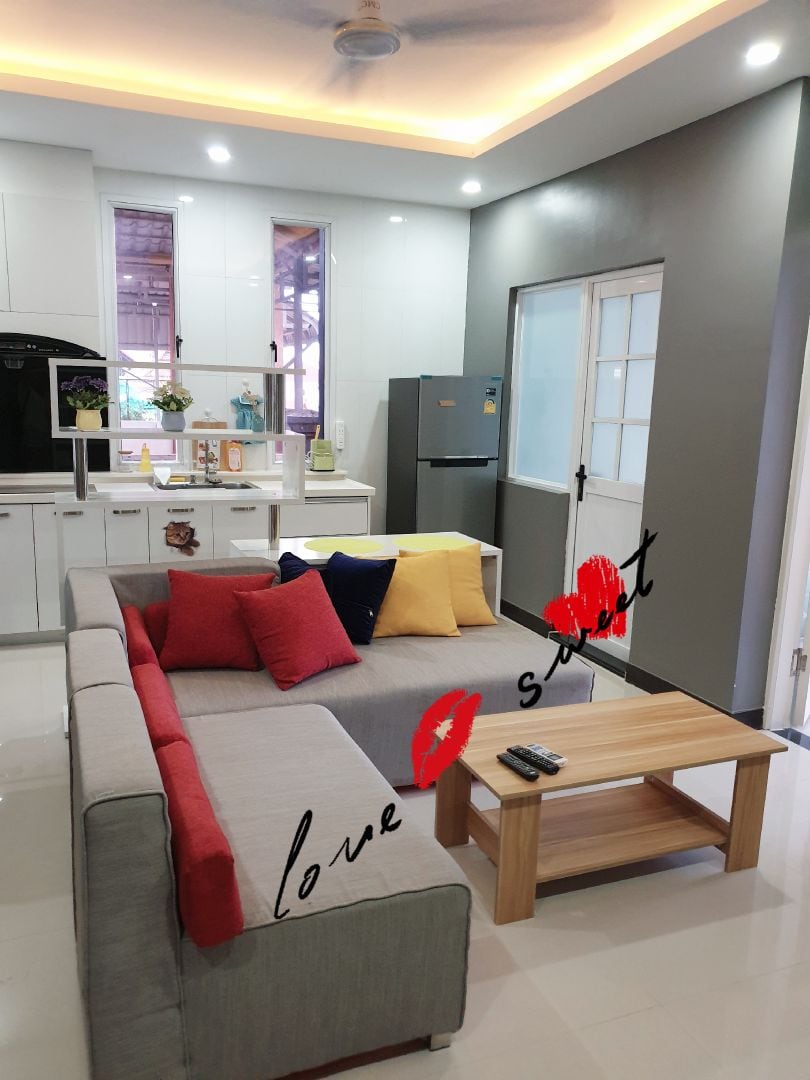 A modern living area features a gray sectional sofa adorned with colorful cushions in red and blue. A light wooden coffee table is central, while a compact kitchen area with white cabinetry and a refrigerator is visible in the background. Bright lighting enhances the space.