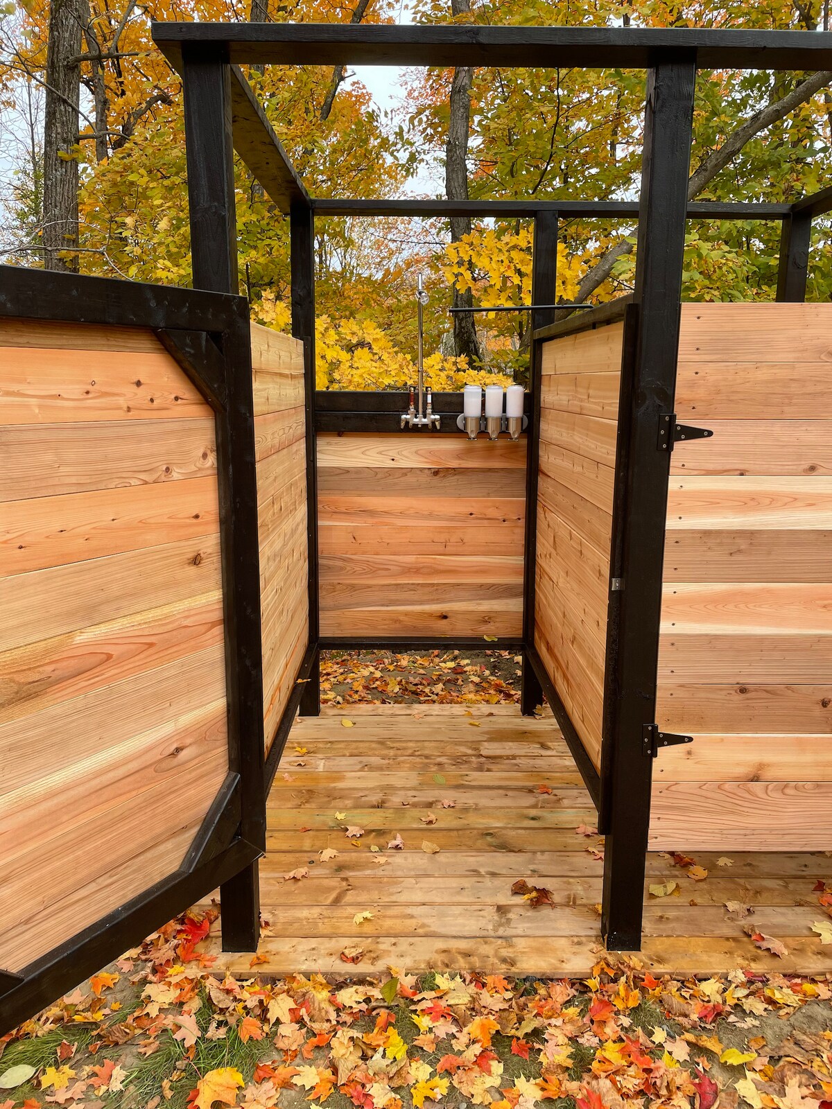 An outdoor shower area is framed by wooden walls, featuring a natural wood floor scattered with colorful autumn leaves. Shower fixtures are mounted on one wall, adding a rustic touch to this private bathing space surrounded by nature.