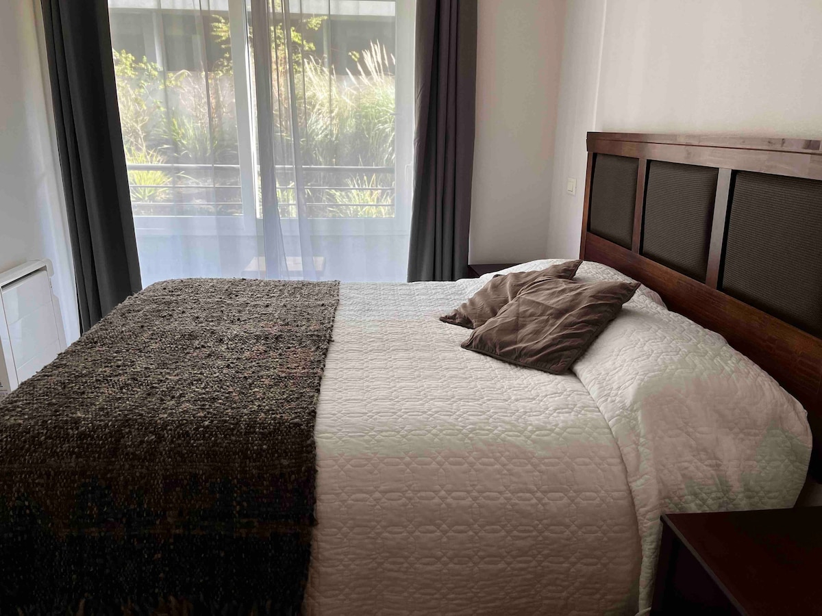 A cozy sleeping area features a spacious bed dressed in a light coverlet, complemented by decorative pillows. Natural light filters through sheer curtains, illuminating the room and revealing views of greenery through the balcony doors.
