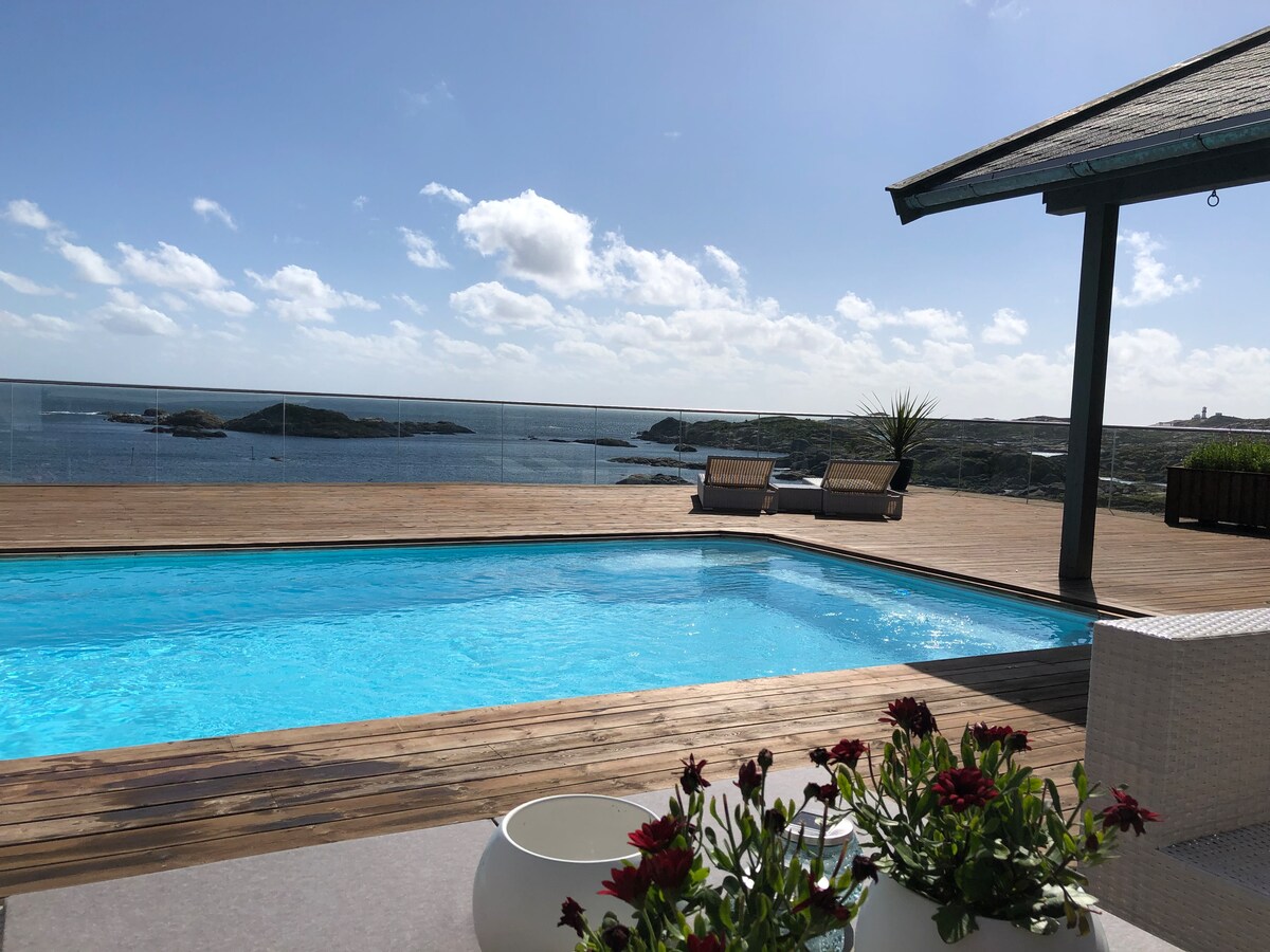 A swimming pool is featured in the foreground, surrounded by a wooden deck. Two loungers are positioned on one side, providing a view of the ocean and distant coastal features under a clear blue sky. Potted flowers add a touch of greenery to the space.