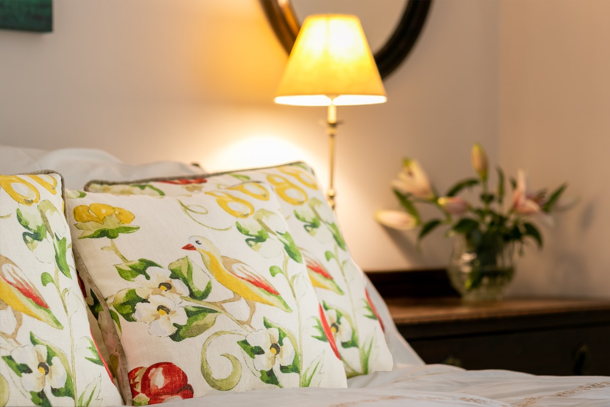 Decorative cushions with floral and bird motifs rest on the bed, complemented by a soft lamp casting a warm glow. A vase of fresh flowers is positioned nearby, contributing to a serene and welcoming ambiance.