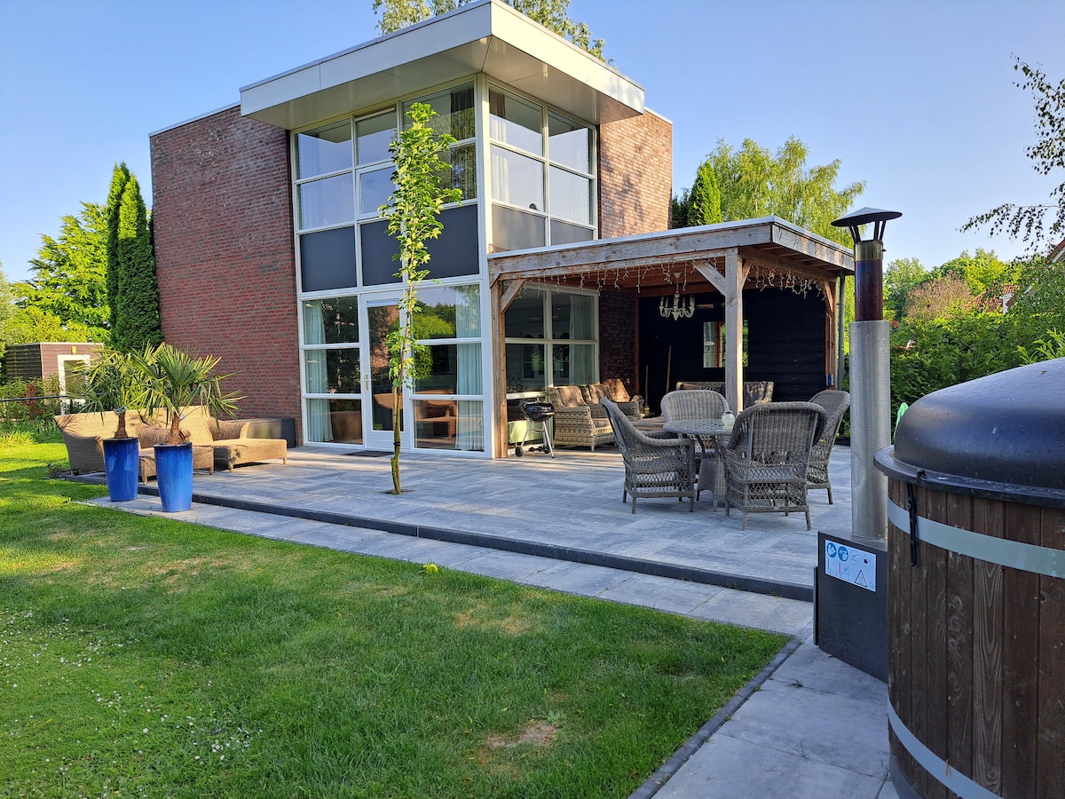 A modern structure features large glass windows and a spacious patio area. Comfortable outdoor seating is arranged under a wooden pergola, accompanied by a dining table. A hot tub is situated nearby, and lush greenery surrounds the space, creating a serene outdoor environment.