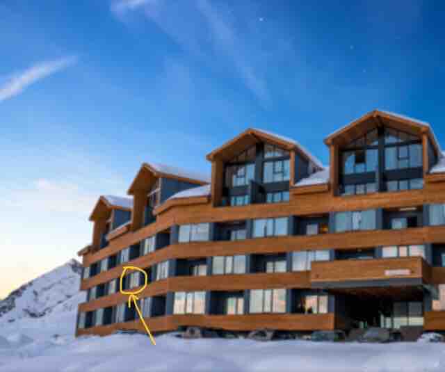 The exterior of a modern apartment building is shown, featuring wooden accents and large glass windows. Snow covers the ground, and the building's upper level has a balcony with a view. A clear sky appears above, enhancing the serene mountain setting.