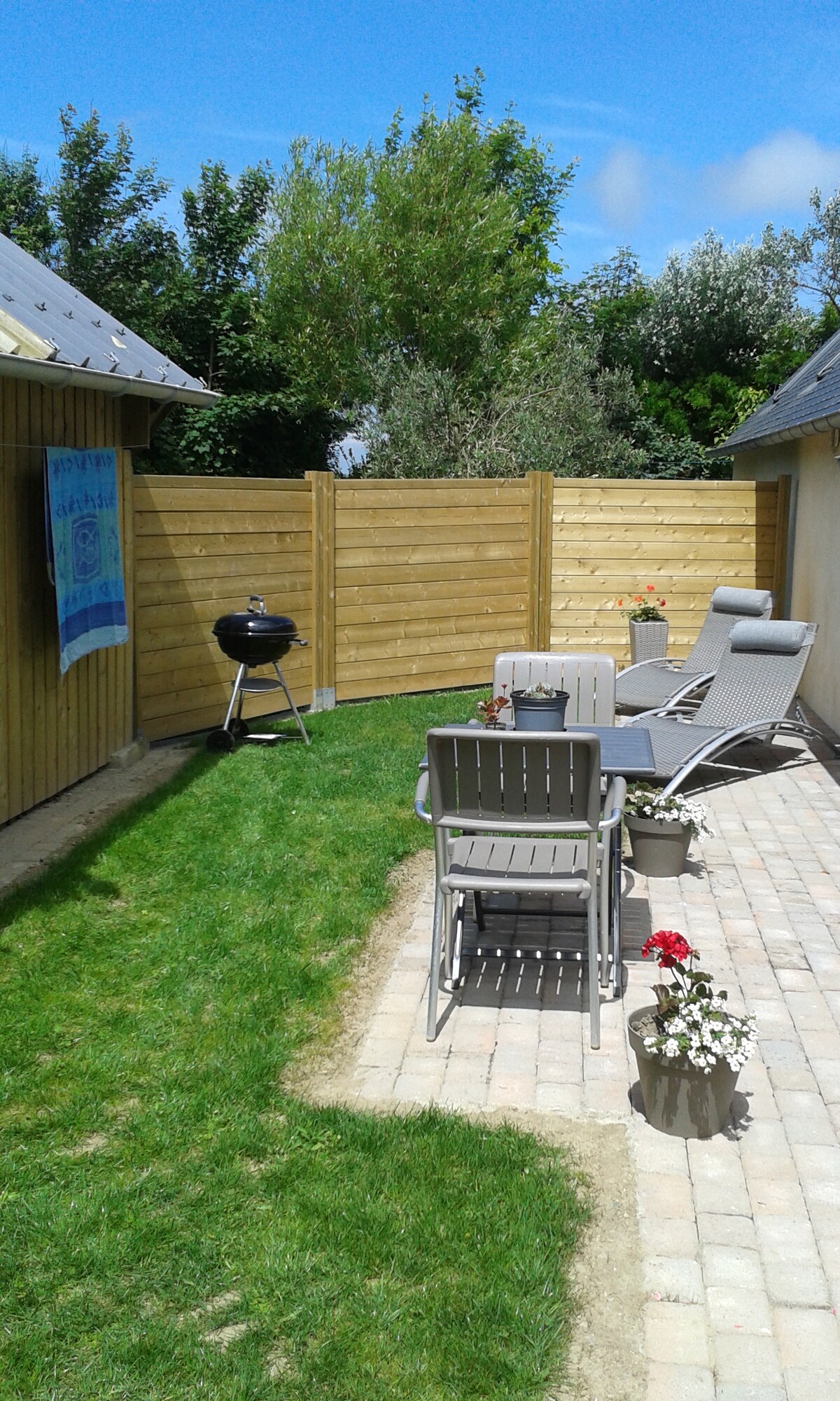 A private outdoor space features a small lawn bordered by wooden fencing. Two lounge chairs and a charcoal grill are positioned on the neatly paved patio, alongside flowering planters, inviting relaxation and outdoor dining.