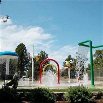 A vibrant water splash pad features colorful structures, including rainbow arches and playful water jets. Surrounding greenery adds a natural touch to the playful environment. The clear blue sky complements the cheerful atmosphere, creating an inviting space for children to enjoy.