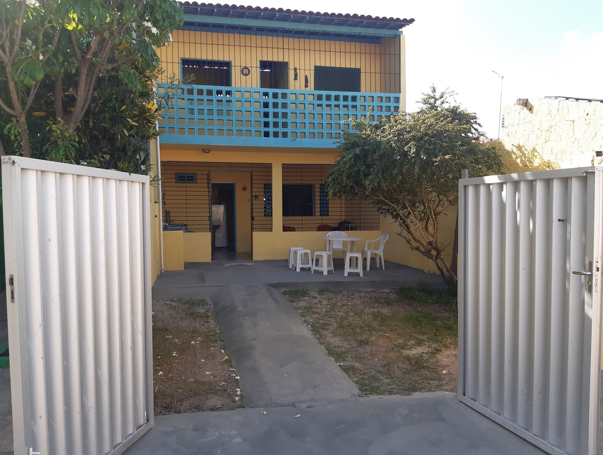 A two-story yellow house is framed by an inviting courtyard, featuring a stone pathway and a small lawn. White outdoor furniture is arranged on a patio area, while the upper balcony is highlighted by a grid-patterned railing, providing a view of the surroundings.