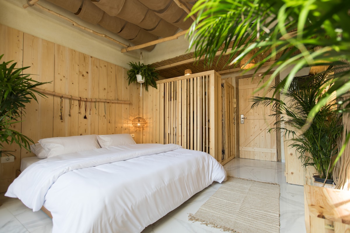 A serene bedroom features a spacious double bed with crisp white linens. Wooden walls and ceiling create a natural atmosphere, while greenery from potted plants adds a touch of nature. The space includes a wooden partition, offering privacy, and warm lighting enhances the inviting ambiance.