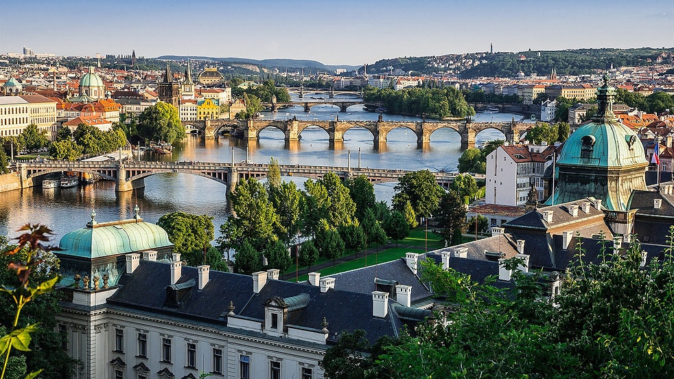 A panoramic view of Prague showcases multiple bridges spanning the Vltava River, surrounded by lush greenery and historic architecture. The scene highlights notable buildings with domed roofs and a vibrant skyline, providing an inviting perspective of the city’s cultural landscape.