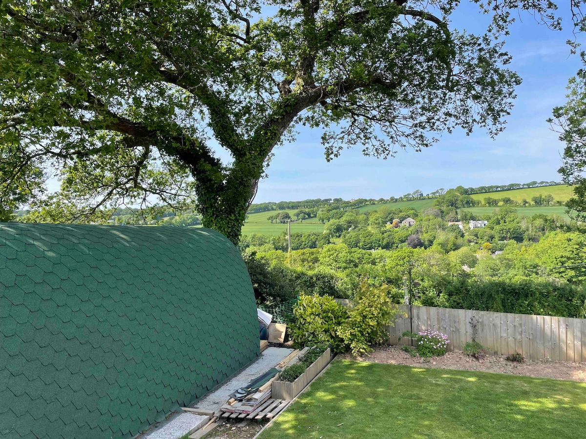A green glamping pod is nestled in a landscaped garden, surrounded by lush greenery. Towering trees provide shade, while scenic hills are visible in the background, showcasing the natural beauty of the Camel Valley. A well-maintained lawn enhances the serene setting.