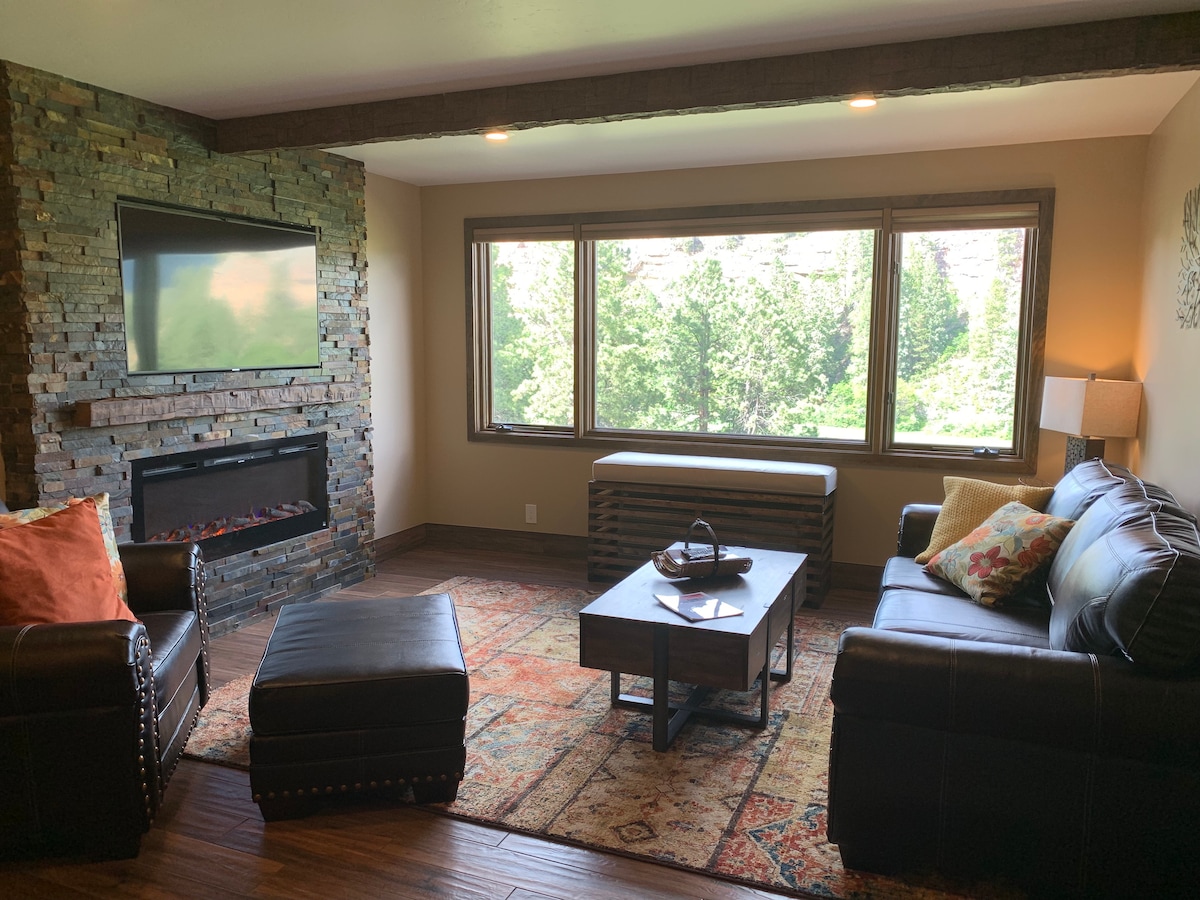 A stylish living room is featured with a stone fireplace and a mounted television. Large windows provide natural light and views of the surrounding greenery. The space is furnished with a dark leather sofa, an ottoman, and an area rug, creating a comfortable atmosphere.