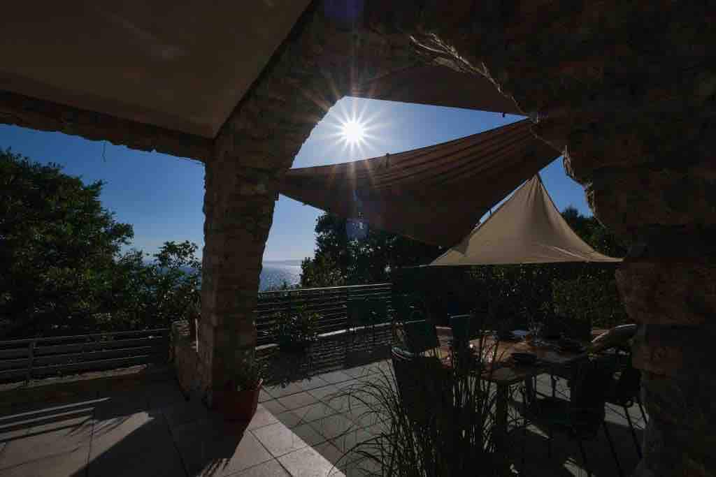 A spacious terrace is shown with stone arches framing a view of clear blue skies and the sun shining brightly. A large table with chairs is positioned beneath a fabric shade sail, while greenery adds a touch of nature to the setting.