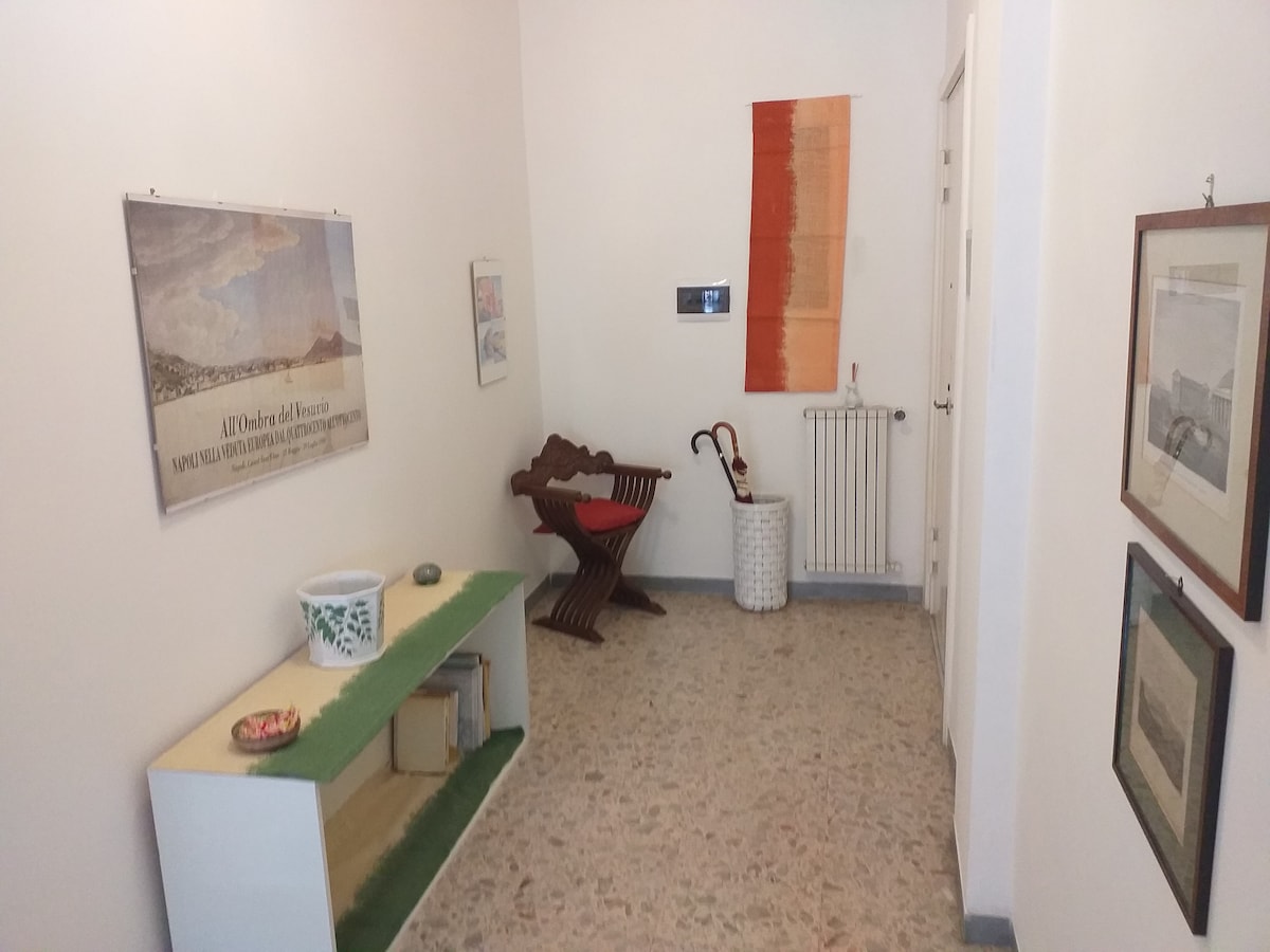 A corridor features light-colored walls adorned with framed artworks and posters. A wooden chair with a unique design is placed against the wall, next to a decorative umbrella holder. A small console table displays potted plants, while a radiator is positioned nearby.