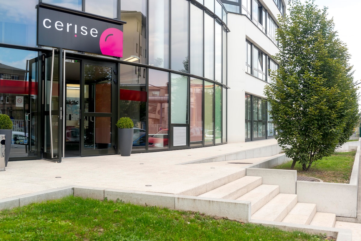 The entrance of La résidence CERISE Strasbourg features a modern façade with large glass windows, allowing natural light to enhance the area. A simple staircase leads up to the entrance, surrounded by well-maintained greenery and decorative planters, creating an inviting approach.
