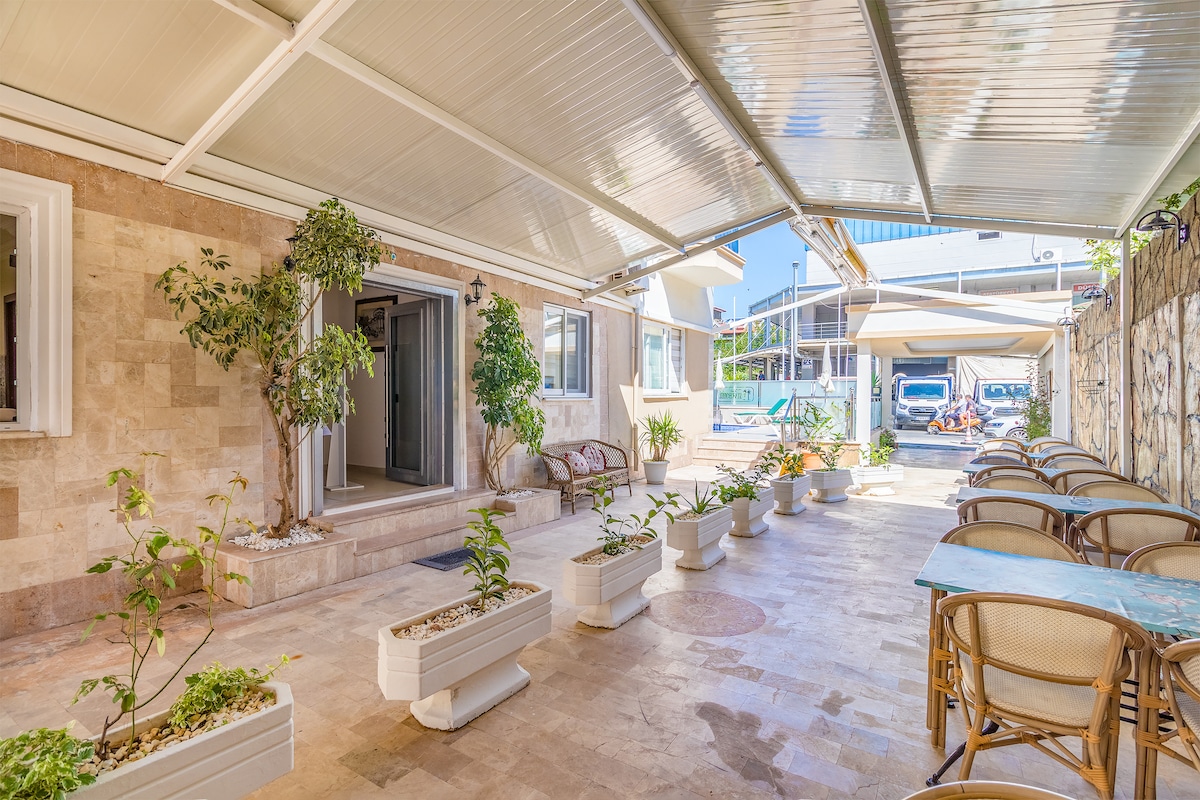A shaded courtyard features a stone floor and potted plants along the pathway. Tables and chairs are arranged for outdoor seating, providing a comfortable area for relaxation. Nearby, an entrance leads into the apartment, offering convenient access to the indoor space.
