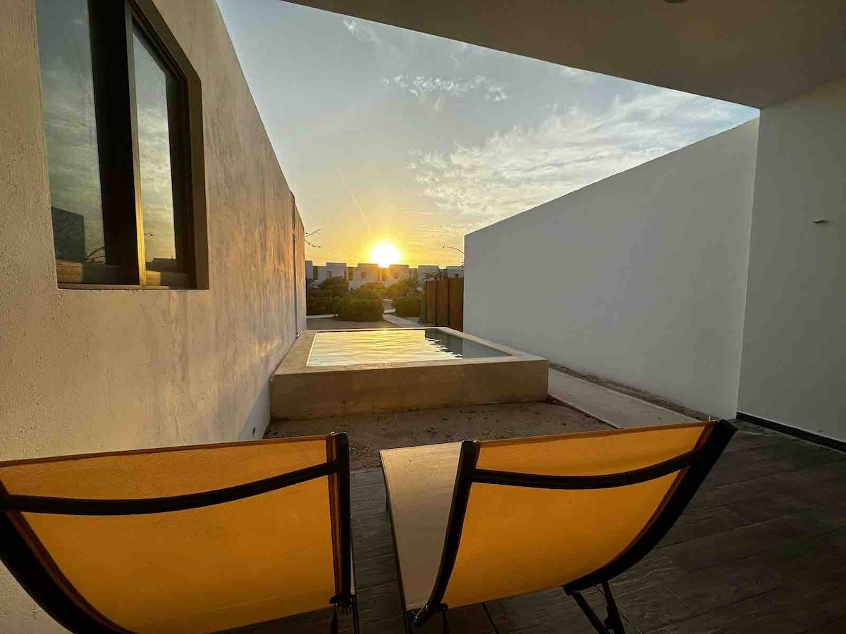 Two yellow chairs are positioned on a wooden deck, facing a serene sunset over a nearby landscape. A small pool reflects the warm light, creating a peaceful ambiance. The surrounding walls are smooth and white, enhancing the open and airy feel of the space.
