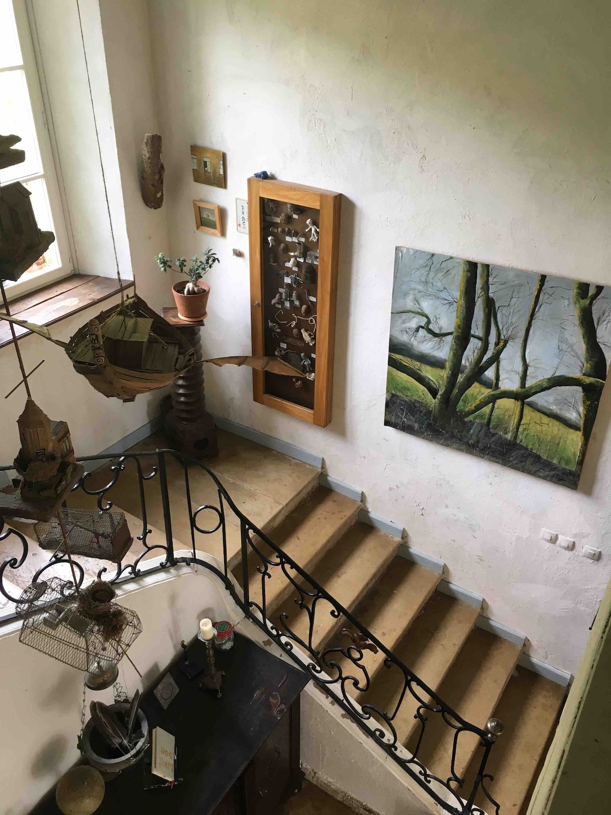 A staircase is visible, adorned with iron railings and flanked by an array of artwork, including a large painting of trees. Natural light enters through a window, illuminating the space. Decorative items and potted plants add charm to the landing.