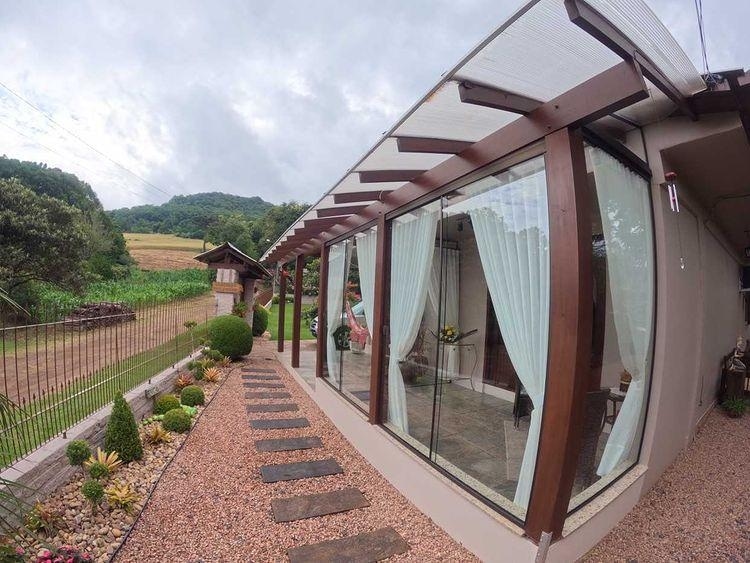 A modern exterior view showcases large glass windows framed in wood, allowing ample light into the space. The pathway is lined with stones, leading to a well-maintained garden filled with greenery. Rolling hills are visible in the background, enhancing the serene natural setting.