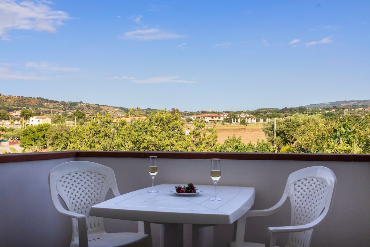 A private terrace offers a view of green hills and distant landscapes. A small white table is set with two glasses and a plate of strawberries, accompanied by two white chairs. The clear blue sky is visible overhead.