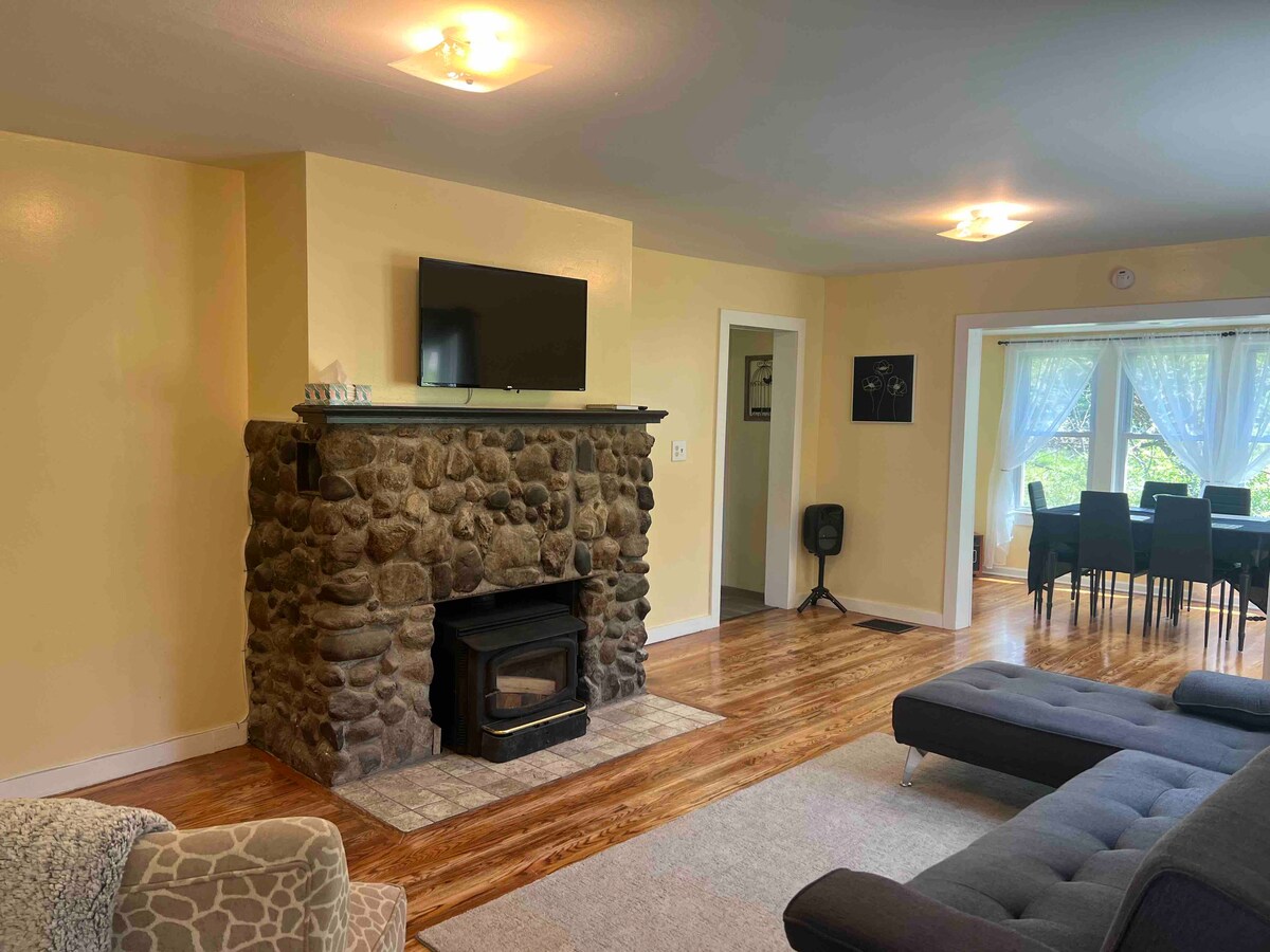 A living room features a rustic stone fireplace with a mounted television above. Natural light filters through sheer curtains, illuminating the room. A dark sectional sofa and a patterned armchair provide comfortable seating. A dining area with a table and chairs is visible in the background.